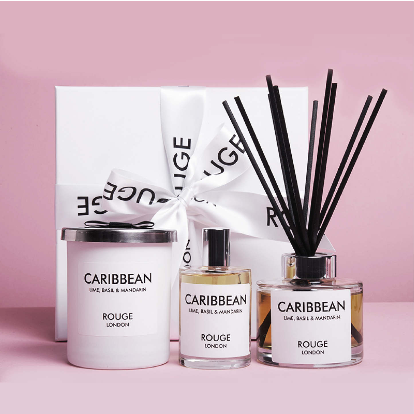 A gift set by Rouge London including a white candle in a container, a reed diffuser and a spray bottle, all labeled 'Caribbean - Lime, Basil & Mandarin'. The items are presented in a white box with a black ribbon.
