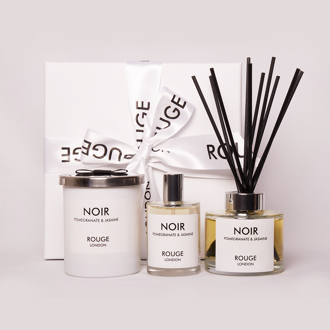 A luxury gift collection set including a white candle in a jar, a reed diffuser with black sticks, and a small spray bottle, all labeled 'NOIR' and 'ROUGE LONDON'. Presented on a two-tone background, light blue and white.